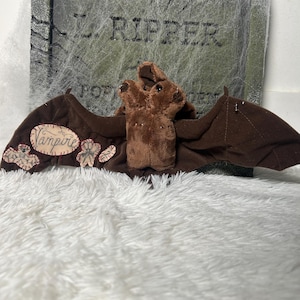 May include: A brown plush bat with outstretched wings, featuring the word "Vampire" on one wing. The bat is set against a backdrop of a tombstone with the name "L. Ripper" and a cobweb design, creating a Halloween theme.