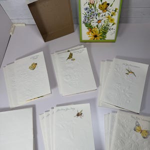 May include: A collection of vintage greeting cards with floral and butterfly designs. The cards are white with embossed floral patterns and feature illustrations of butterflies, hummingbirds, and bees. A green box with a floral illustration is also present.