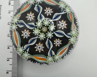 Puede incluir: Un pisapapeles redondo con fondo negro y diseño floral colorido. El diseño incluye elementos blancos, verdes, naranjas y azules. El pisapapeles mide aproximadamente 5 cm de diámetro.