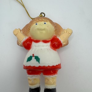 Puede incluir: Adorno navideño vintage con forma de muñeca. La muñeca tiene pelo rubio, ojos azules y viste un atuendo rojo y blanco con zapatos negros. Se adjunta un cordón dorado para colgar.