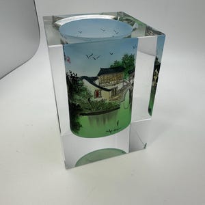 May include: A clear, square glass candle holder with a cylindrical insert featuring a painted landscape scene. The scene depicts a traditional building with a bridge over a green body of water, under a blue sky.