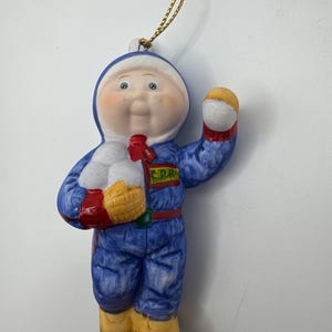 Può includere: Ornamento in ceramica raffigurante un bambino in tuta da neve blu con cappuccio bianco. L'ornamento ha una sciarpa rossa, stivali gialli e un guanto giallo e rosso. Il bambino tiene in mano un oggetto giallo. È attaccato un cordino dorato per appenderlo.