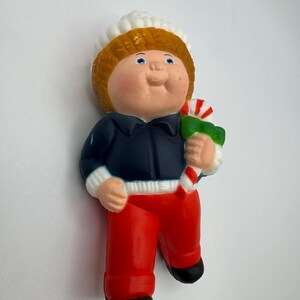 May include: A vintage figurine of a child wearing a white hat, a navy blue jacket, and red pants. The figurine is holding a green and red candy cane. The child has orange hair and rosy cheeks. The figurine's shoes are black.