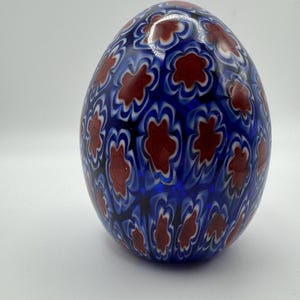 May include: A decorative glass egg with a vibrant blue base color. The egg is adorned with a repeating pattern of red flower-like shapes, outlined in white, creating a striking visual contrast. The egg is a unique decorative item.