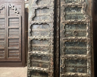 May include: A pair of tall, weathered wooden doors featuring a dark brown and teal patina. The doors have a rectangular panel design with arched top sections, decorative metal studs, and a circular metal ring handle centred on the right panel.