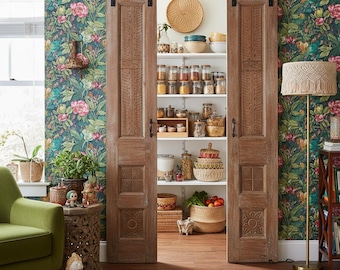 May include: A rustic pantry with sliding wooden doors. The doors are open, revealing shelves filled with jars, baskets, and bowls. The walls are covered in a floral wallpaper. A green armchair and a floor lamp with a macrame shade are visible.
