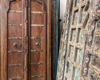 Könnte beinhalten: Ein Set aus hohen, rechteckigen Holztüren mit aufwendig geschnitzten Rändern und metallenen Ringgriffen. Die Türen haben eine satte braune Oberfläche mit sichtbarer Holzmaserung, während angrenzende Paneele eine verwitterte, abgenutzte Textur mit blauer Farbe aufweisen.