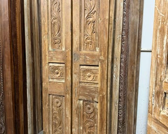 May include: Two weathered wooden doors with carved floral and scrollwork designs. The doors are a light brown color and have a rectangular shape. Other doors with similar designs are stacked in the background.