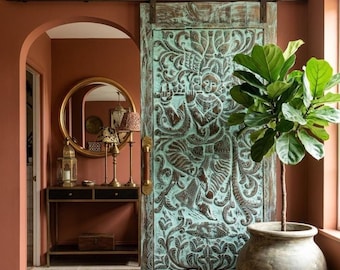 May include: A sliding barn door featuring a teal and brown finish with intricate hand-carved relief patterns depicting a figure playing a flute, mounted on a dark metal track system with a vertical brass handle, positioned against a terracotta wall.