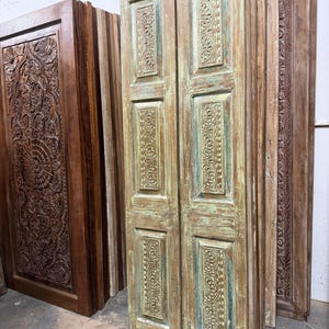 May include: A collection of antique wooden doors with intricate carved floral and geometric designs. The doors are various shades of brown and green, with some showing signs of age and wear. The doors are stacked together, showcasing their unique textures and patterns.