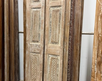 May include: Aged wooden doors with intricate carvings and a distressed finish. The doors feature rectangular panels with geometric and floral designs. The wood has a natural, weathered appearance, suggesting a vintage or antique style. The doors are leaning against a wall.