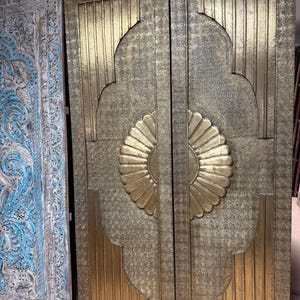 May include: Two ornate, golden doors with intricate patterns and a central sunburst design. The doors are made of a metallic material and feature vertical and curved lines, creating a textured appearance. A light blue carved wooden door is visible on the left.