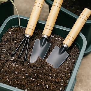 Könnte beinhalten: Drei Gartengeräte mit Holzgriffen und schwarzen Metallköpfen. Die Geräte befinden sich in einem grünen Pflanztopf, der mit dunkelbrauner Erde gefüllt ist.