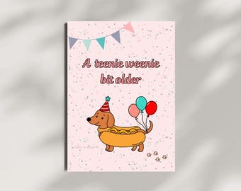 May include: A pink birthday card featuring a cartoon dachshund in a hot dog bun, wearing a party hat and holding balloons. The text reads "A teenie weenie bit older" in pink script, with paw prints at the bottom.