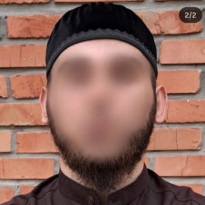 May include: A black velvet kufi hat with a decorative trim. The hat is worn by a person with a dark beard and is set against a brick wall background.