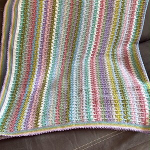 May include: A rectangular crochet blanket featuring a repeating vertical striped pattern in pastel shades of lavender, mint green, soft pink, light blue, and yellow, finished with a textured border in matching colors and a scalloped edge.