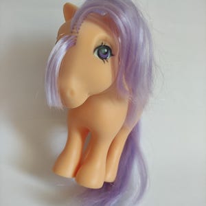 Può includere: Un giocattolo vintage a forma di pony con un corpo color pesca e capelli lavanda. Il pony ha grandi occhi dettagliati e una criniera e una coda fluenti. Il giocattolo è un oggetto da collezione classico.