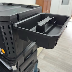 May include: A black modular tool storage system featuring a stackable design with a removable, open-top plastic tray extended outward. The tray includes a central handle and internal dividers, while the main unit has side ventilation slots.
