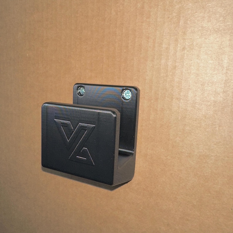以下が含まれることがあります： VZの文字がエンボス加工された長方形のフロントプレートを備えたマットブラックの壁掛けフック。フックはU字型のプロファイルで、上部の角にある2本の目に見える銀色の金属ネジで表面に固定されています。