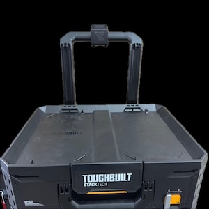 Può includere: Una cassetta degli attrezzi nera ToughBuilt StackTech dotata di un vano portaoggetti rettangolare con maniglia superiore. La parte anteriore mostra la scritta bianca ToughBuilt StackTech e un'etichetta IP65. Il design include un telaio robusto.