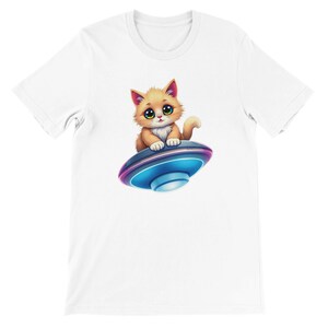 Puede incluir: Una camiseta blanca con una imagen de dibujos animados de un lindo gato naranja montando un platillo volador. El gato tiene grandes ojos azules y una nariz rosa.