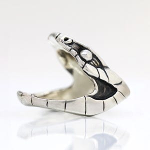 Könnte beinhalten: Ein silberner Ring in Form eines Schlangenkopfes mit offenem Maul. Der Ring hat detaillierte Schuppen und schwarze Akzente an Maul und Augen. Er wird auf einer weißen Oberfläche mit Spiegelung gezeigt.