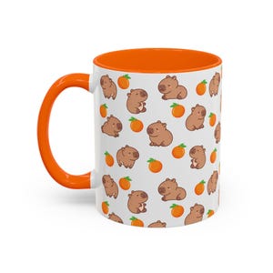 Könnte beinhalten: Weiße Keramiktasse mit orangefarbenem Rand und einem Muster aus braunen Capybaras und orangen Zitrusfrüchten.