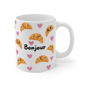 Könnte beinhalten: Weiße Keramiktasse mit einer Schwarzweißillustration von Croissants mit Gesichtern und rosa Herzen. Der Text "Bonjour" ist auf die Tasse gedruckt.