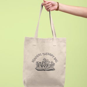 May include: A beige tote bag featuring the text "Readers Therapy Bag" above an illustration of an open book and flowers. The bag is held by a person against a light green backdrop.