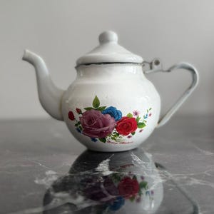 Pode incluir: Bule de esmalte branco com um design floral de rosas vermelhas, roxas e azuis. O bule tem uma alça curva e um bico. A tampa tem um pequeno botão. O bule está sobre uma superfície reflexiva.