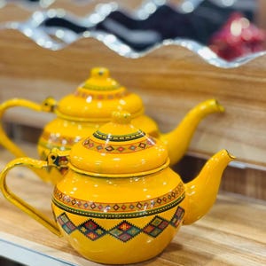 Può includere: Due teiere gialle con manici e beccucci decorativi. Le teiere hanno coperchi e sono decorate con motivi geometrici colorati attorno alla base e sui coperchi. Le teiere sono su una superficie di legno.