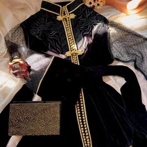 May include: A black velvet jacket with gold detailing and decorative clasps. The jacket has sheer, dotted sleeves and is shown with a matching clutch. A perfume bottle and decorative vase are also present.