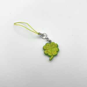 May include: A four-leaf clover charm in shades of green and yellow, attached to a yellow cord and a silver-colored clasp. The clover is a symbol of good luck and is a popular accessory.