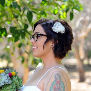 May include: A bride with short, dark hair wears black-framed glasses and a white flower in her hair. She is wearing a white strapless dress and has a tattoo of the Virgin Mary on her shoulder.