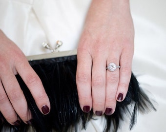 May include: A woman's hand with dark red nail polish is wearing a silver ring with a large, round, clear diamond. The ring is on the ring finger of the right hand. The hand is holding a black feather clutch.