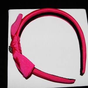 May include: A pink satin headband with a black elastic band and a pink satin bow with a silver accent.