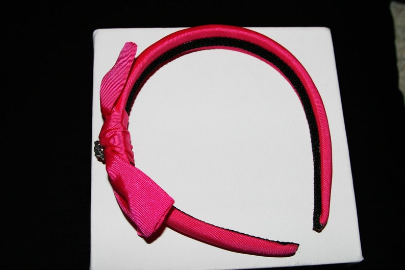 May include: A pink satin headband with a black elastic band and a pink satin bow with a silver accent.