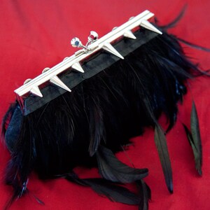 May include: A black feather clutch with a silver metal frame and spikes. The clutch is closed and has a small, silver, decorative detail on the front.