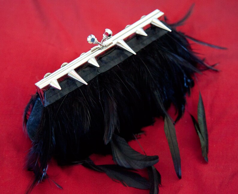 May include: A black feather clutch with a silver metal frame and spikes. The clutch is closed and has a small, silver, decorative detail on the front.