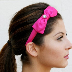 May include: A pink satin headband with a rhinestone bow. The headband is worn by a person with long brown hair pulled back in a ponytail.