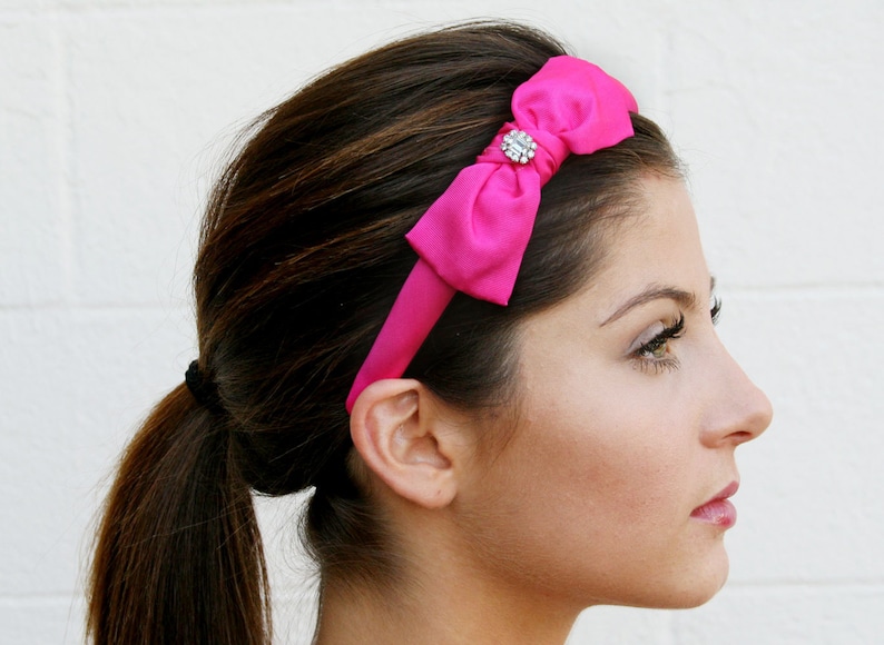 May include: A pink satin headband with a rhinestone bow. The headband is worn by a person with long brown hair pulled back in a ponytail.