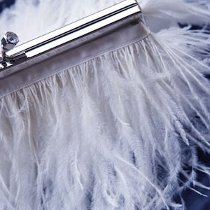 Puede incluir: Un bolso de mano blanco con cierre plateado y flecos de plumas blancas.