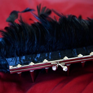 May include: A black feather clutch with a silver metal frame and pointed studs. The clutch has a silver clasp with a clear rhinestone.