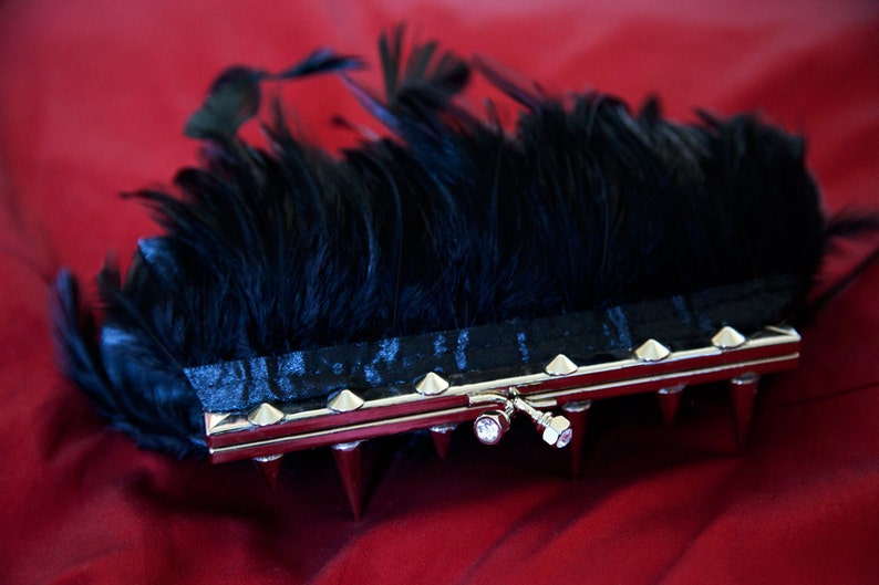 May include: A black feather clutch with a silver metal frame and pointed studs. The clutch has a silver clasp with a clear rhinestone.