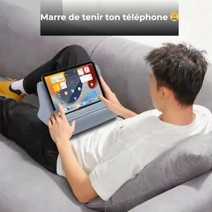Puede incluir: Una persona usa una tableta con un soporte azul claro mientras está reclinada en un sofá gris. La tableta muestra una pantalla de inicio colorida con iconos de aplicaciones. El texto en la parte superior dice "Marre de tenir ton téléphone".
