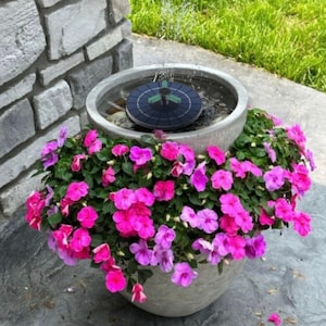 May include: A solar-powered fountain in a large, grey concrete planter filled with vibrant pink and purple flowers. The fountain is circular, with a dark panel and small water jets, creating a small water display. The planter sits on a grey stone surface.