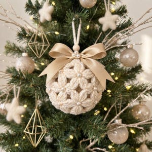 May include: A round, cream-coloured crocheted ornament featuring a floral pattern with small white beads at the centre of each flower. The ornament is topped with a smooth, light beige satin ribbon bow and a small wooden bead for hanging.