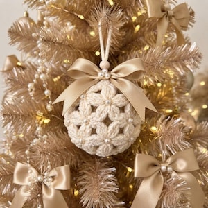 May include: A round, cream-colored crocheted ornament featuring a floral pattern with small white beads at the center of each flower. The ornament is topped with a smooth, light beige satin ribbon bow and hangs from a matching cream-colored cord.