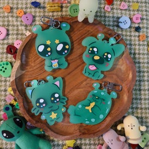 May include: A collection of green alien-themed keychains with various designs, including one holding a drink. The keychains are on a wooden tray, surrounded by colorful buttons and small alien figurines. The background has a patterned fabric.