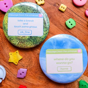May include: Two round pinback buttons featuring computer-style pop-up windows. One button has a green grass background with the text "message from the universe", "take a break and touch some grass", and "ok, fine". The other is blue with "message from the universe", "where do you wanna go?", and "home".